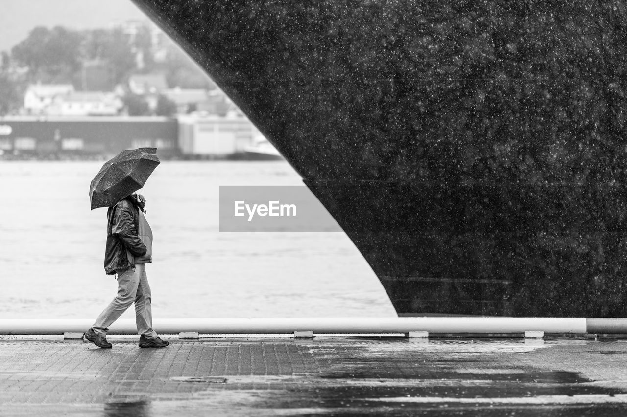 Man walking with umbrella on wet sidewalk by cruise ship during monsoon