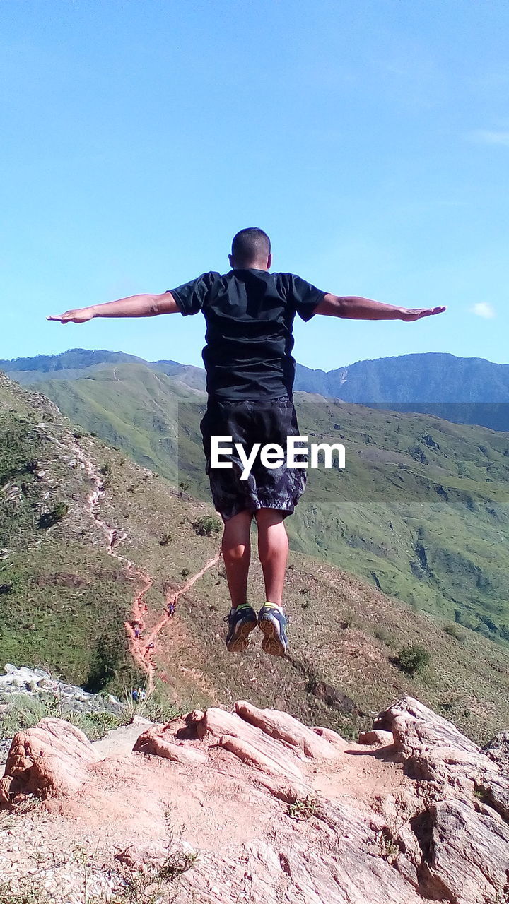 Rear view of man with arms outstretched over rock at mountains against sky