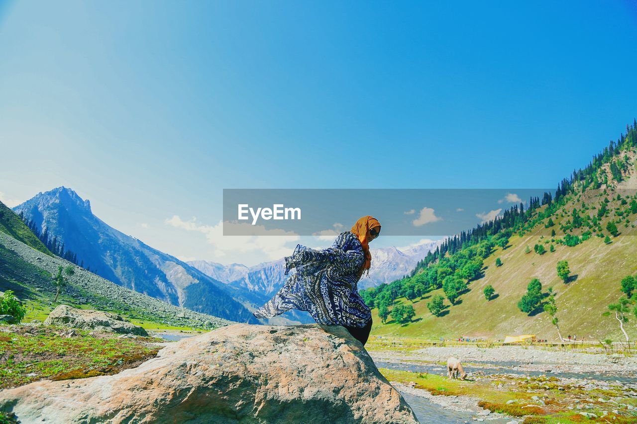 Side view of woman sitting on rock against sky