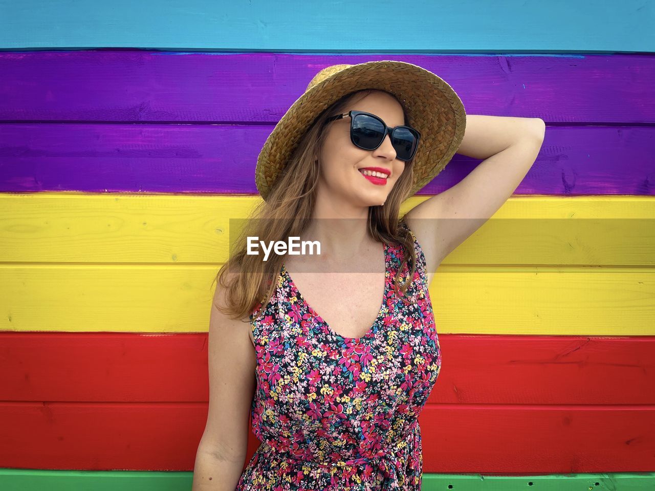 Fashionable young woman wearing straw hat and sunglasses standing against bright and colorful wall