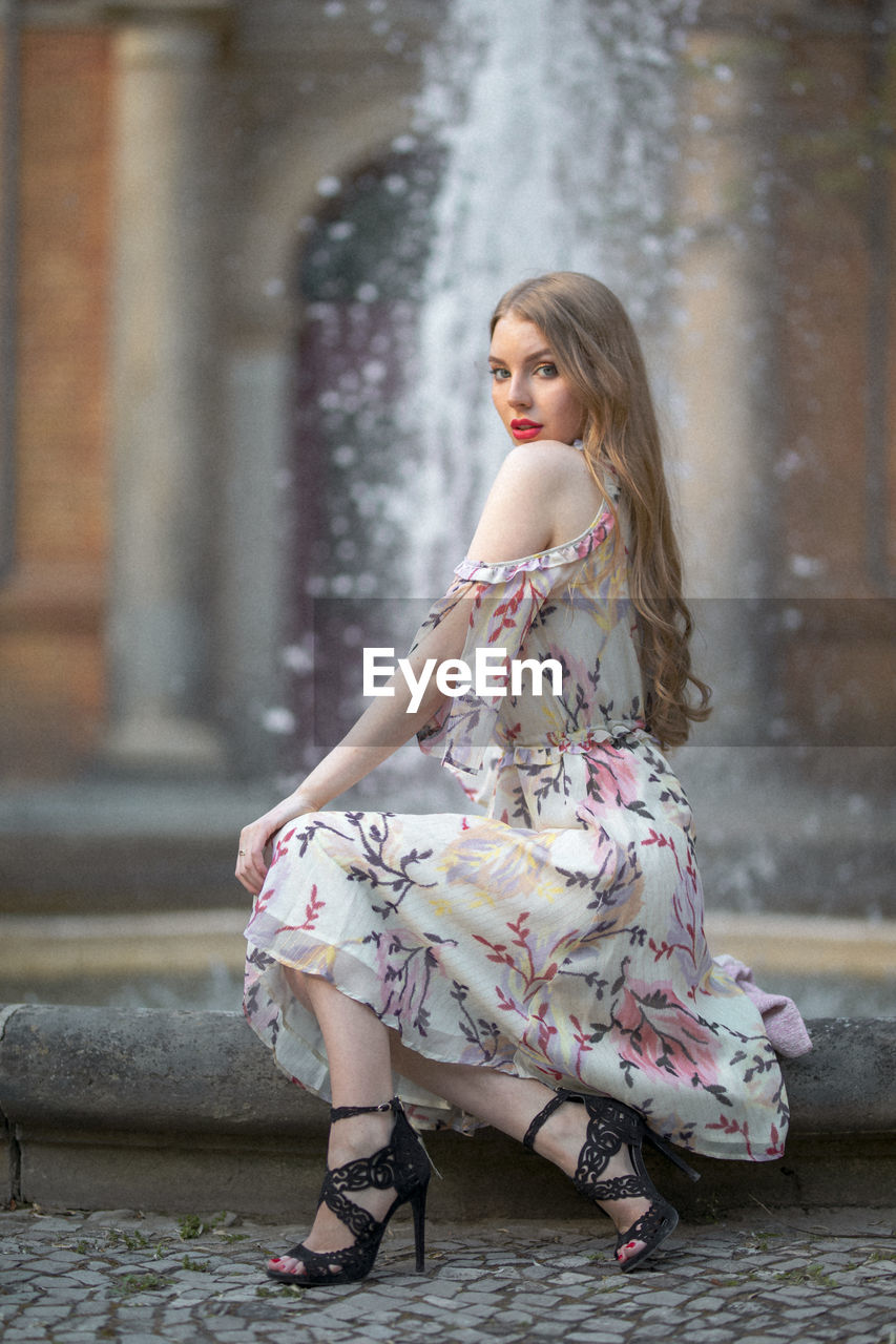 Portrait of beautiful young woman sitting against fountain in city