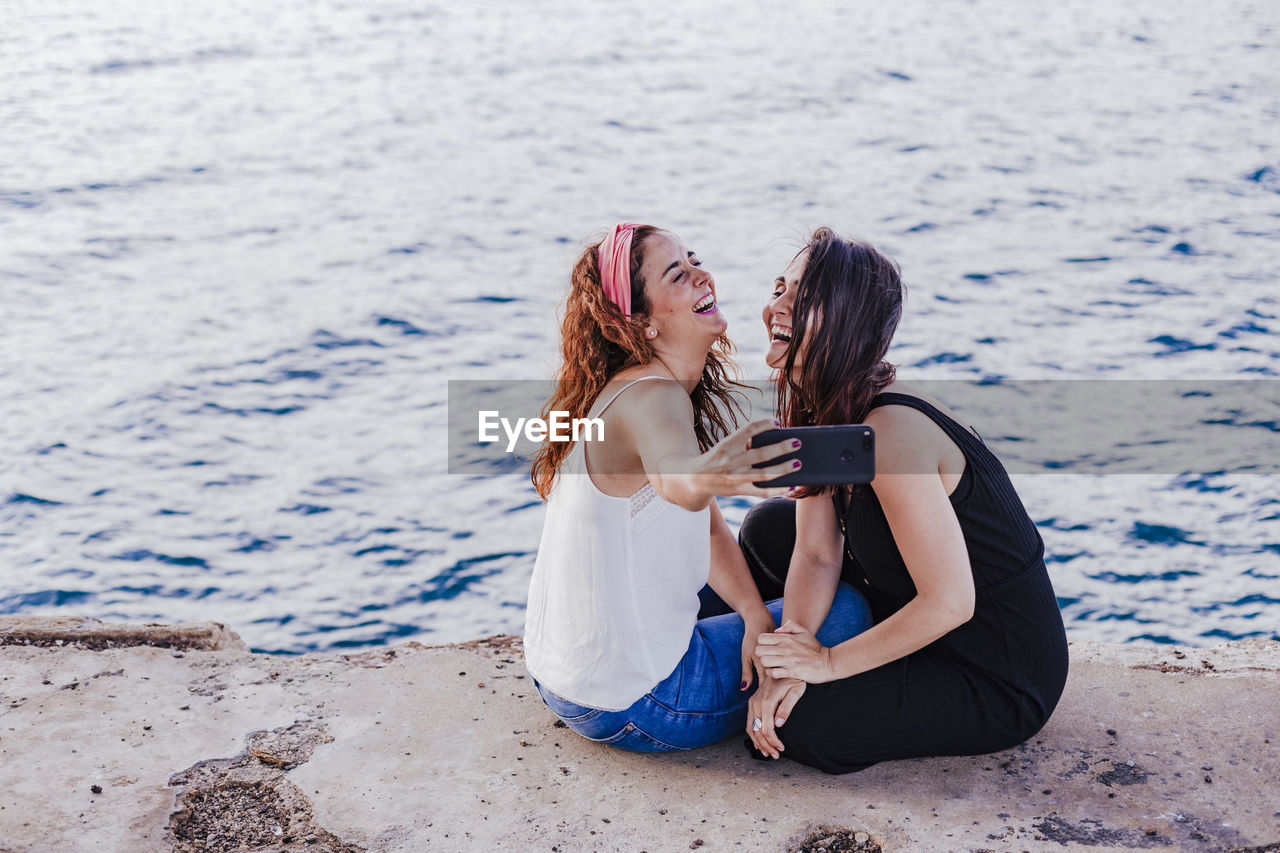 Lesbian couple taking selfie while sitting by sea