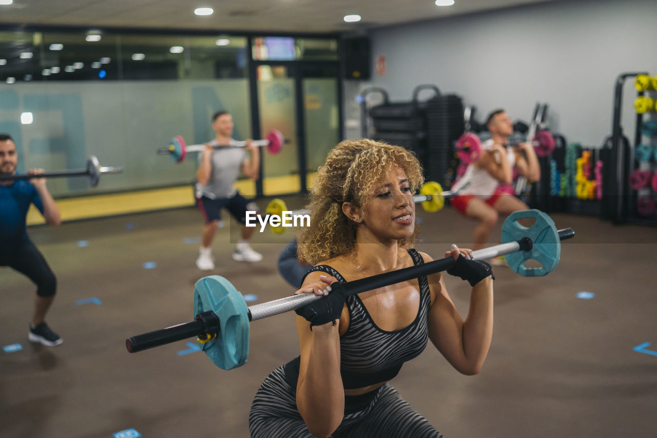 Curly haired female athlete lifting barbell while exercising with friends in gym