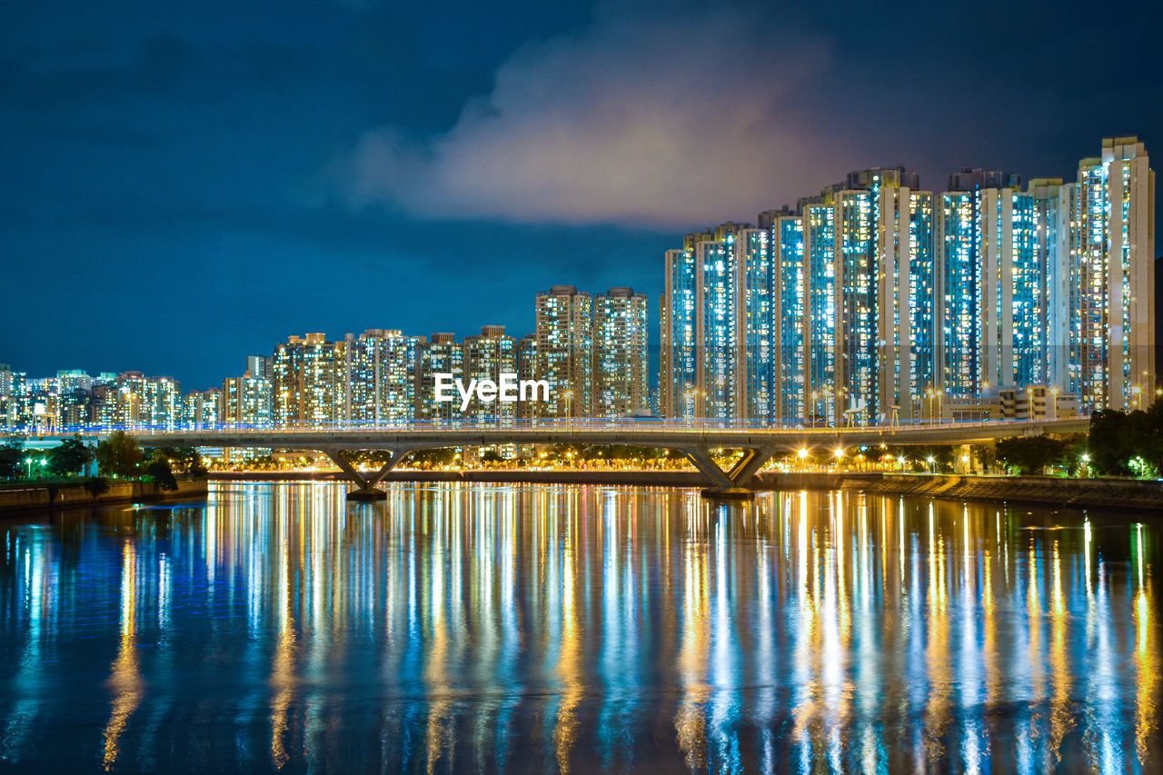 Illuminated modern buildings by river against sky at night