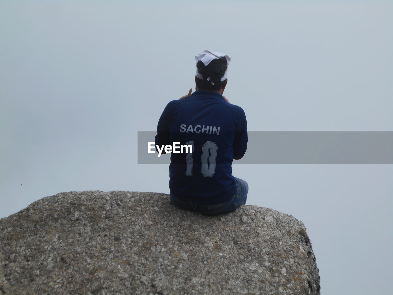 REAR VIEW OF MAN SITTING ON ROCK AGAINST SKY