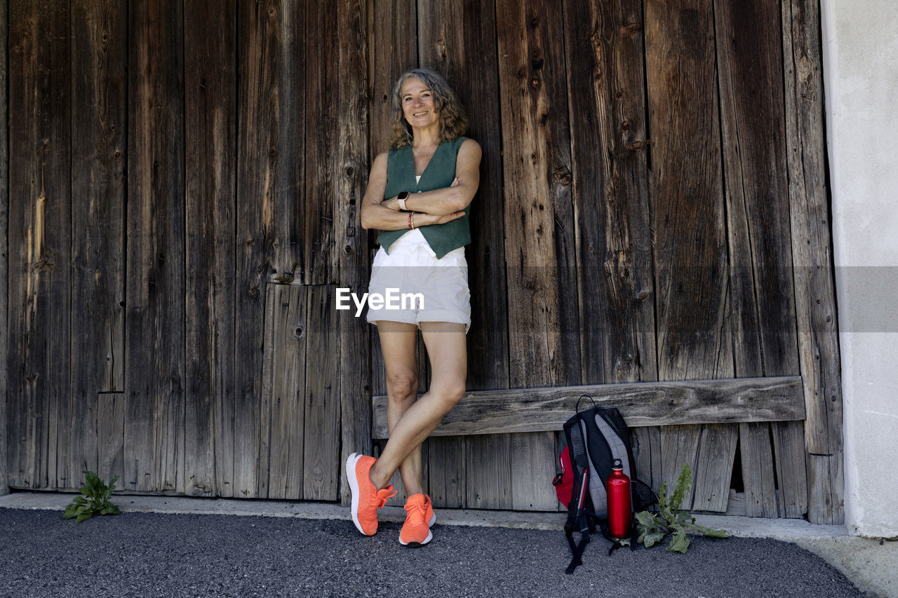 Happy senior woman wearing orange shoes with arms crossed leaning on old wooden barn door