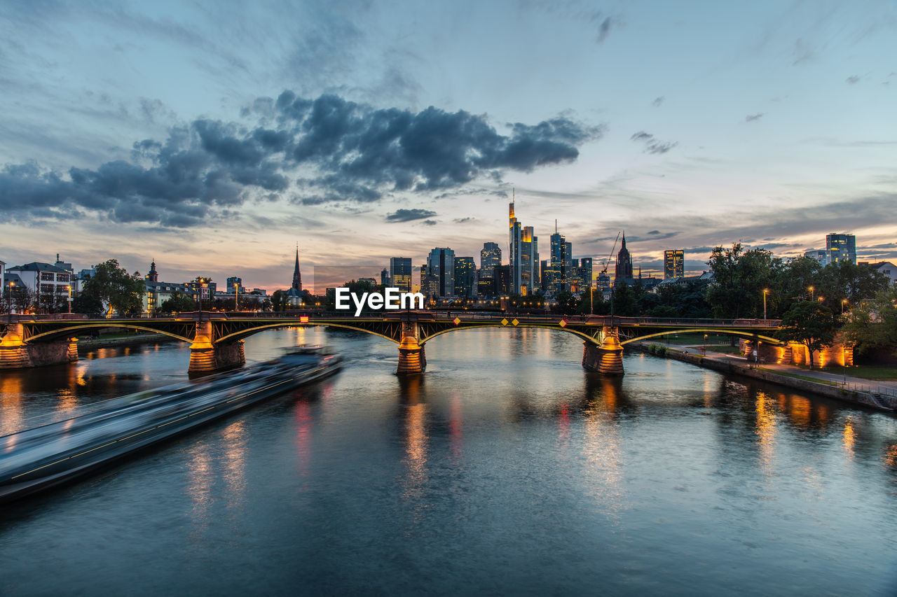 BRIDGE OVER RIVER BY ILLUMINATED BUILDINGS AGAINST SKY