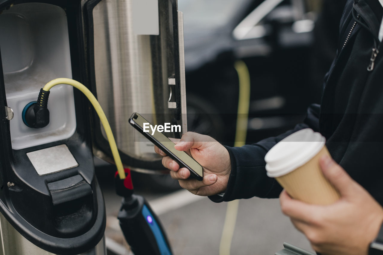 Midsection of man using smart phone at electric car charging station