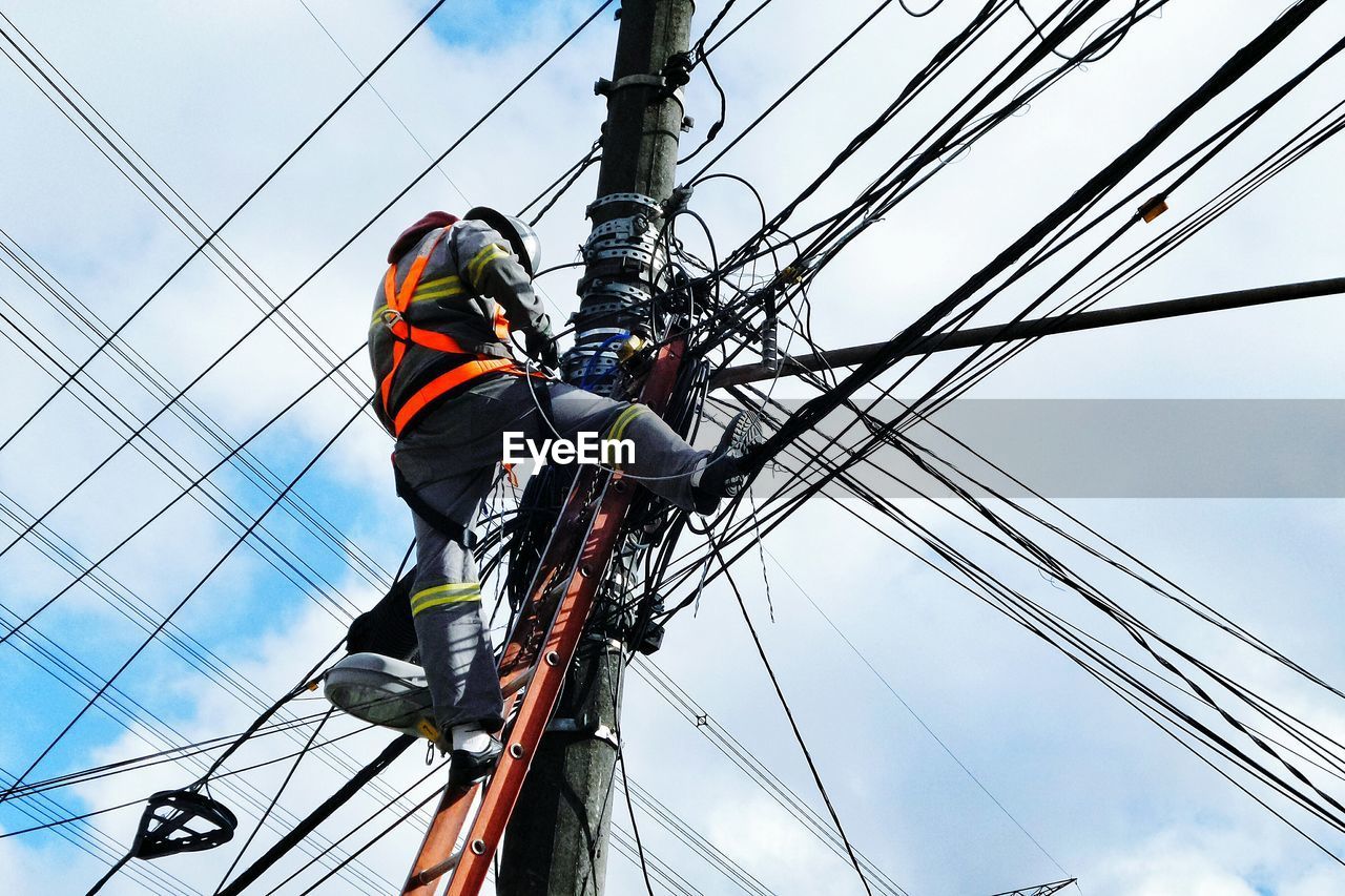 Low angle view of electrician working on pole against cloudy sky