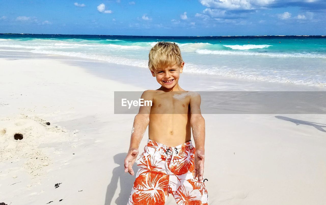 Portrait of shirtless boy standing at beach against sky