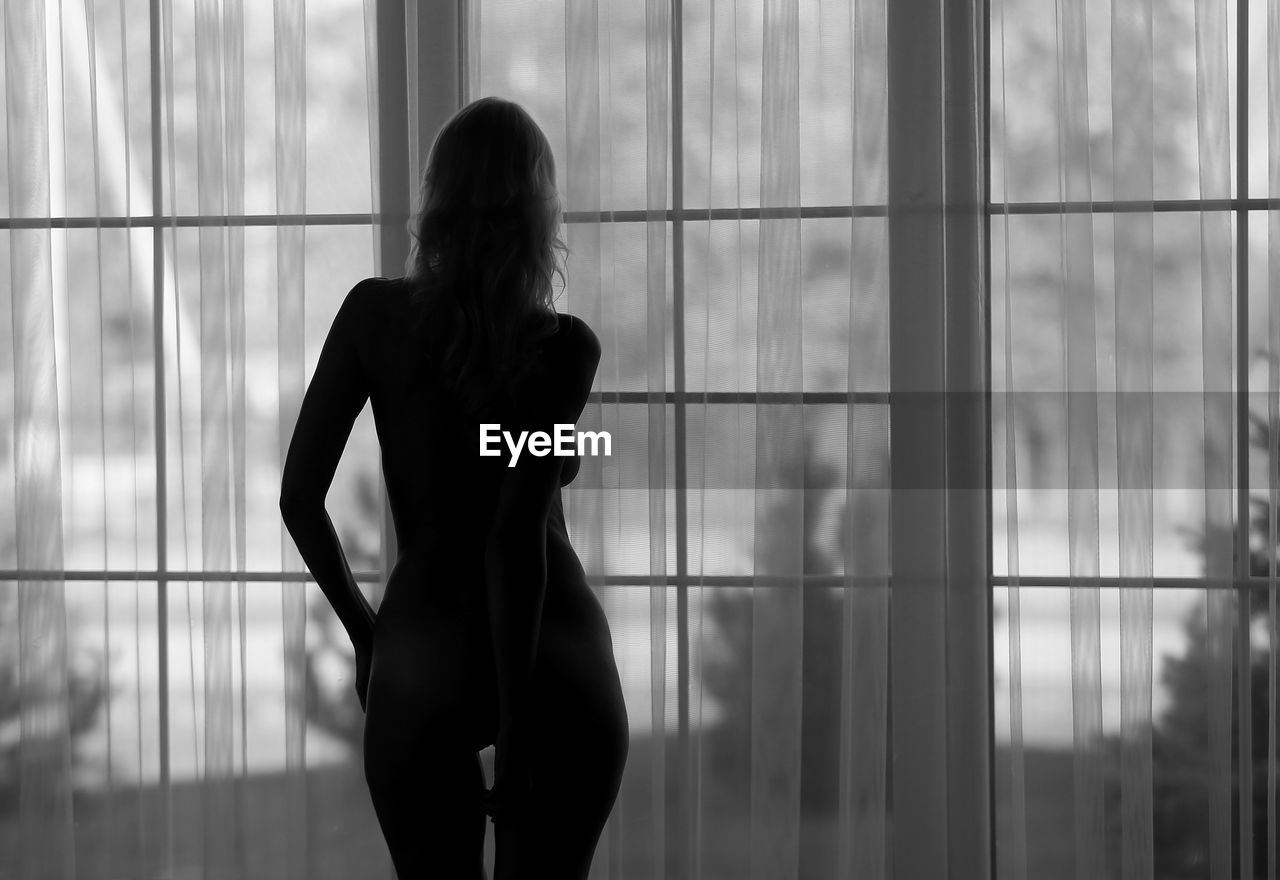 Rear view of naked woman standing by window at home 