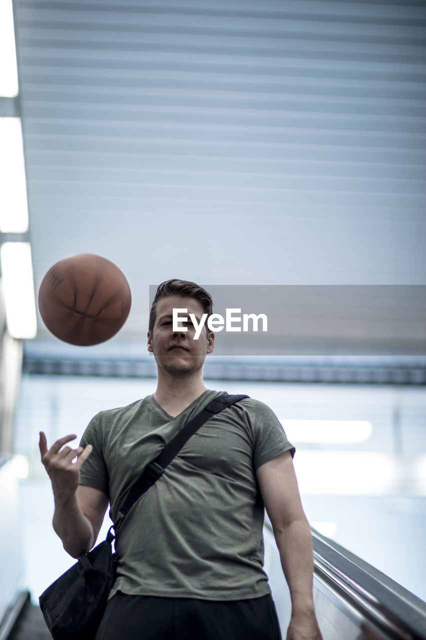 Low angle portrait of young man playing with basketball while moving down on escalator