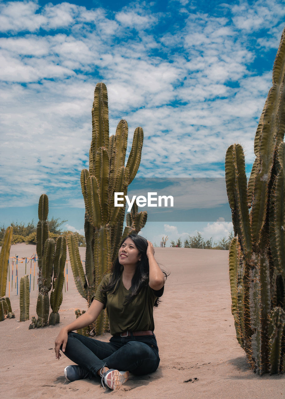 Full length of smiling woman sitting by cactus in desert