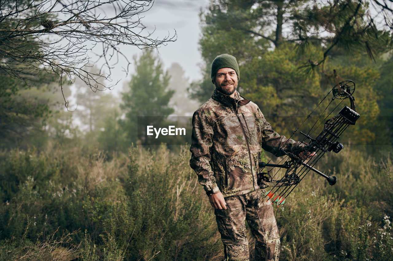 Cheerful man in camouflage standing with compound bow in forest and looking at camera during hunting