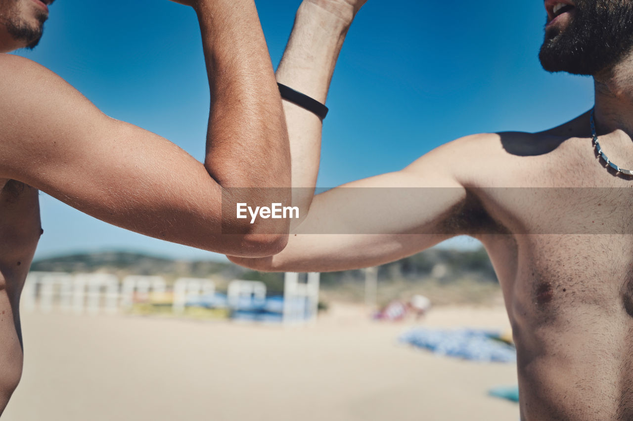 Midsection of shirtless men following social distancing with elbow bump at beach