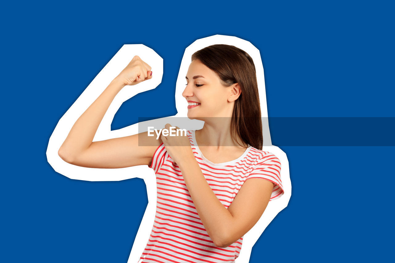 Smiling young woman flexing muscles against blue background