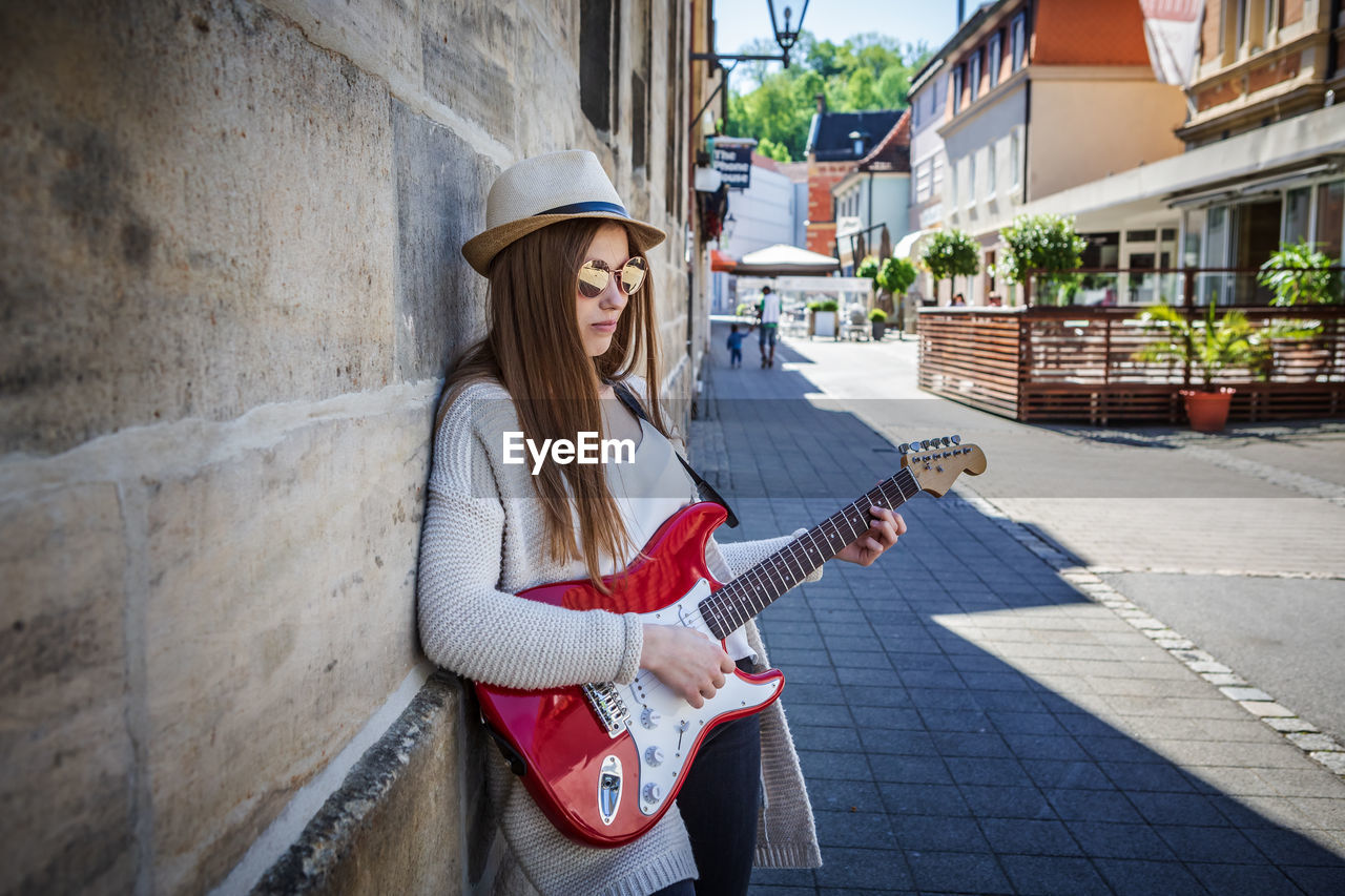 Young woman playing guitar while standing by wall