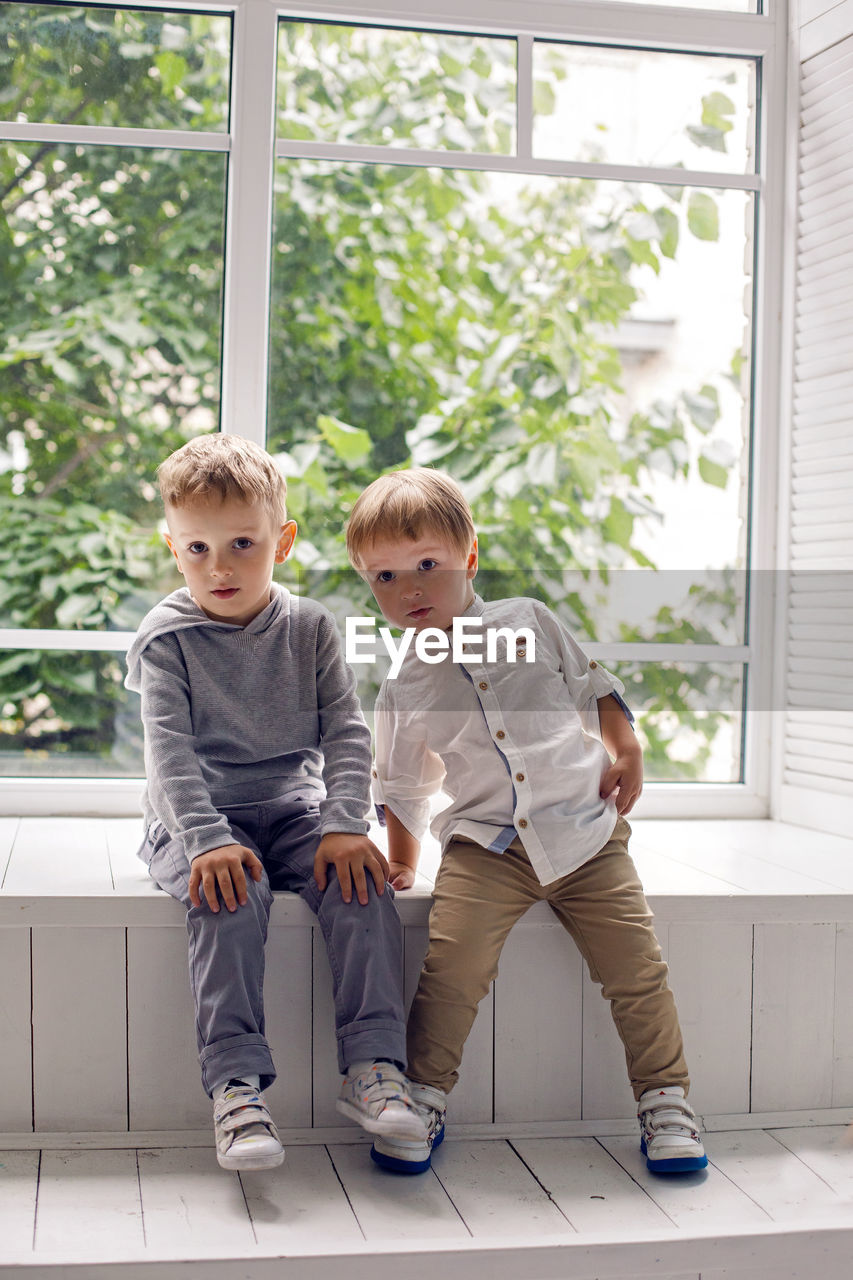 Two brothers sitting by the window in a white studio with green trees on the street