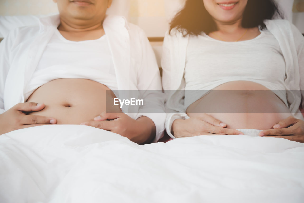 Midsection of pregnant woman with husband sitting on bed at home