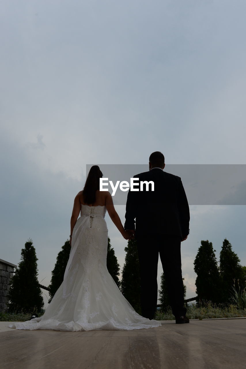 Rear view of bride and groom standing on walkway against cloudy sky