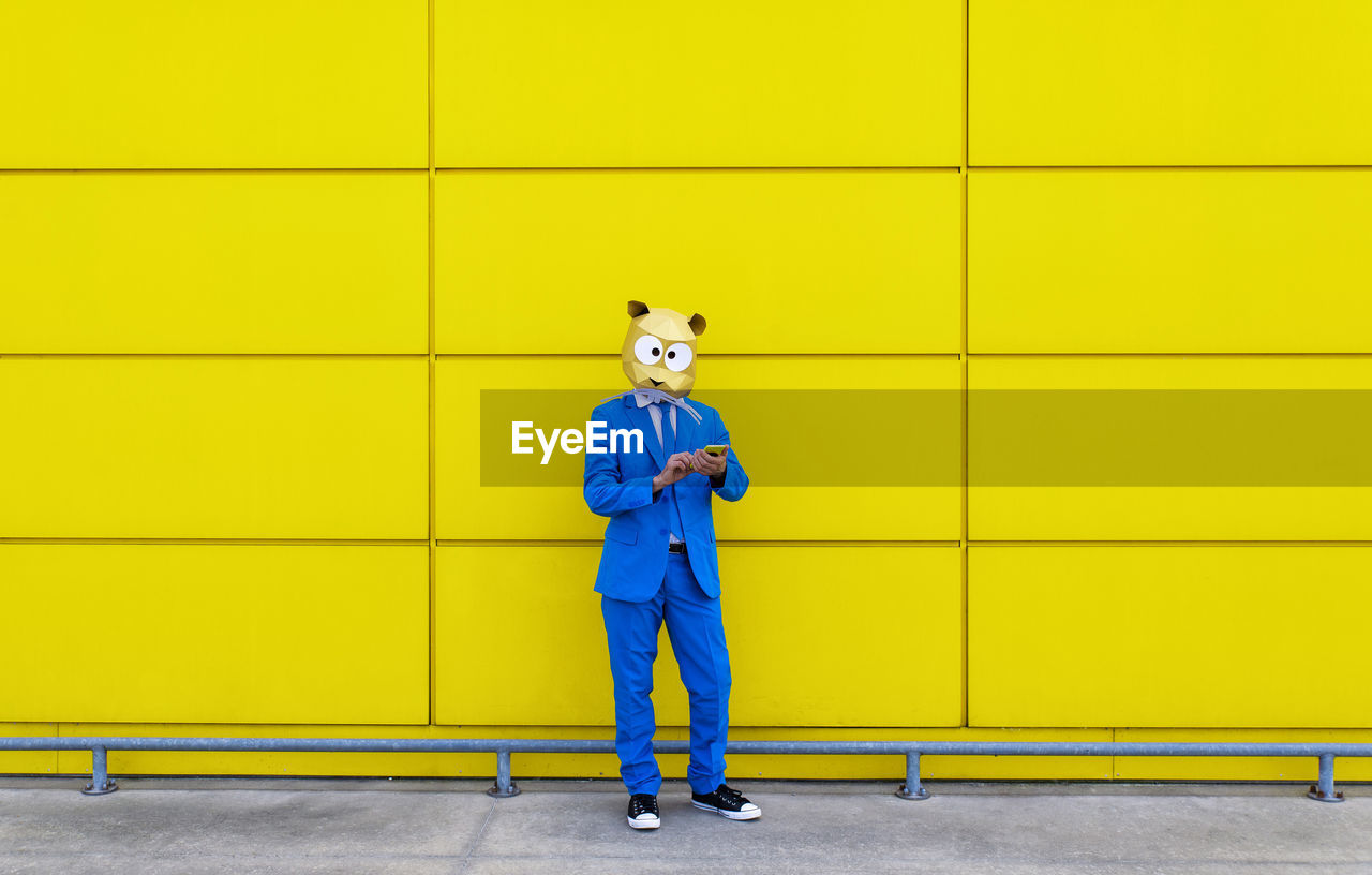 Man wearing vibrant blue suit and rodent mask standing in front of yellow wall with smart phone in hands