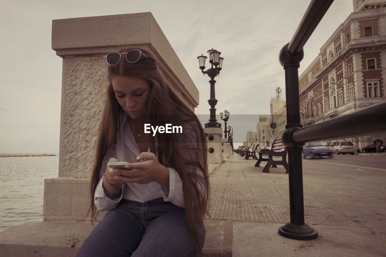 Young woman using phone while sitting in city against sky