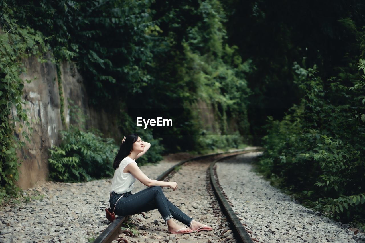 Side view of woman sitting on railroad track amidst trees in forest