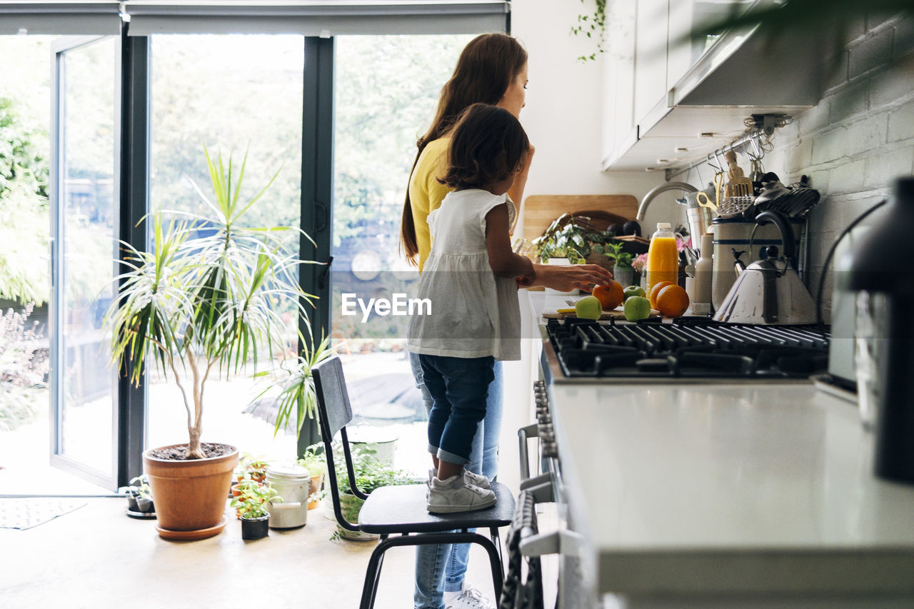 Girl standing on chair by woman preparing food in kitchen