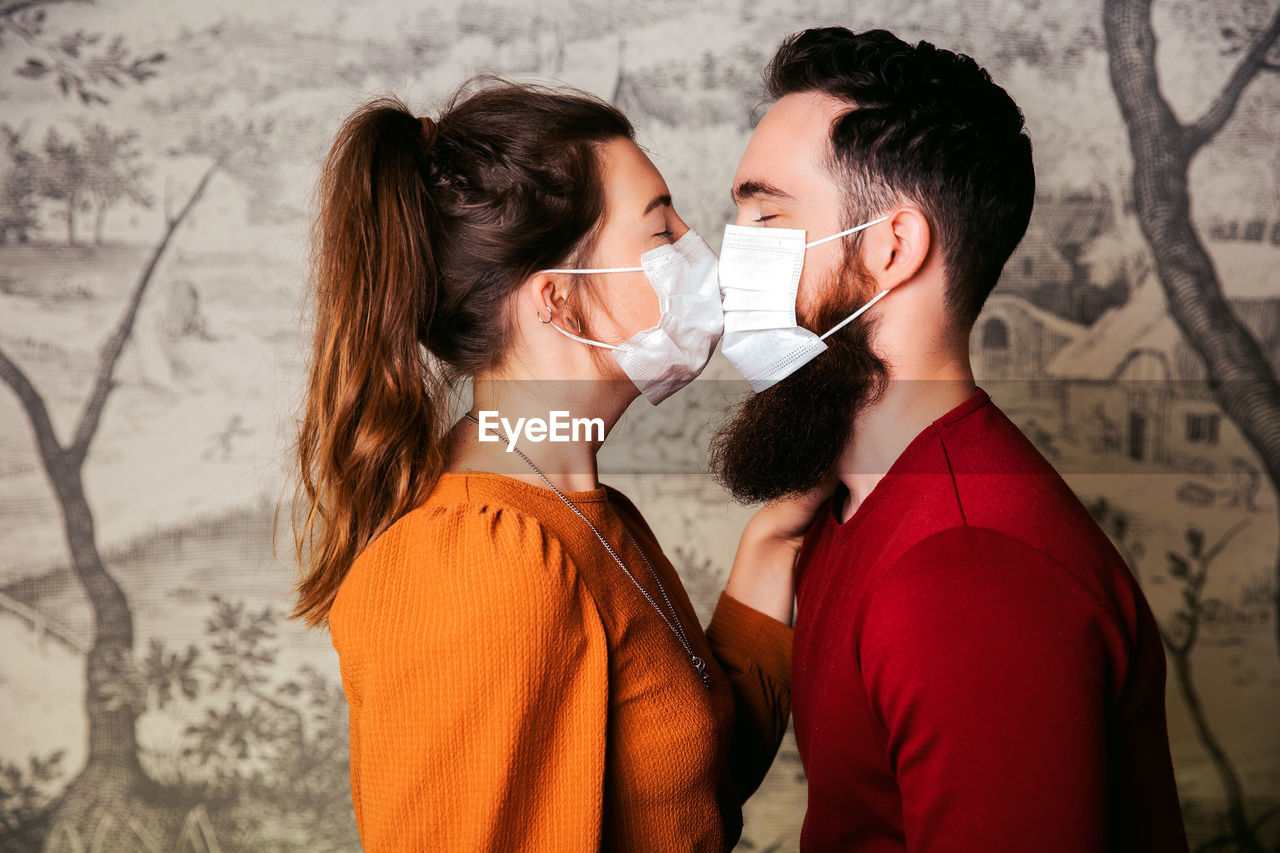 Side view of young couple wearing masks kissing against wall