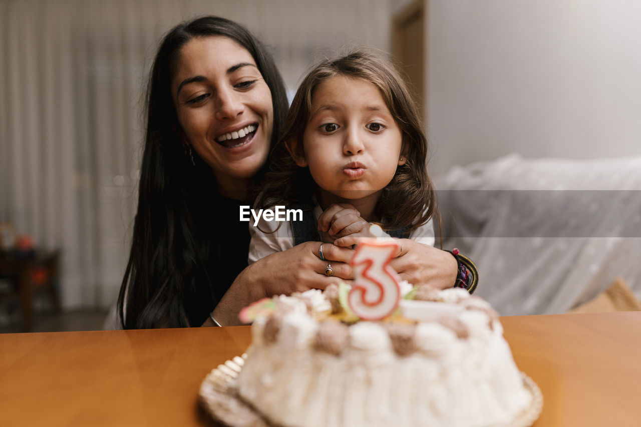 Girl blowing candle over birthday cake while sitting with mother at home