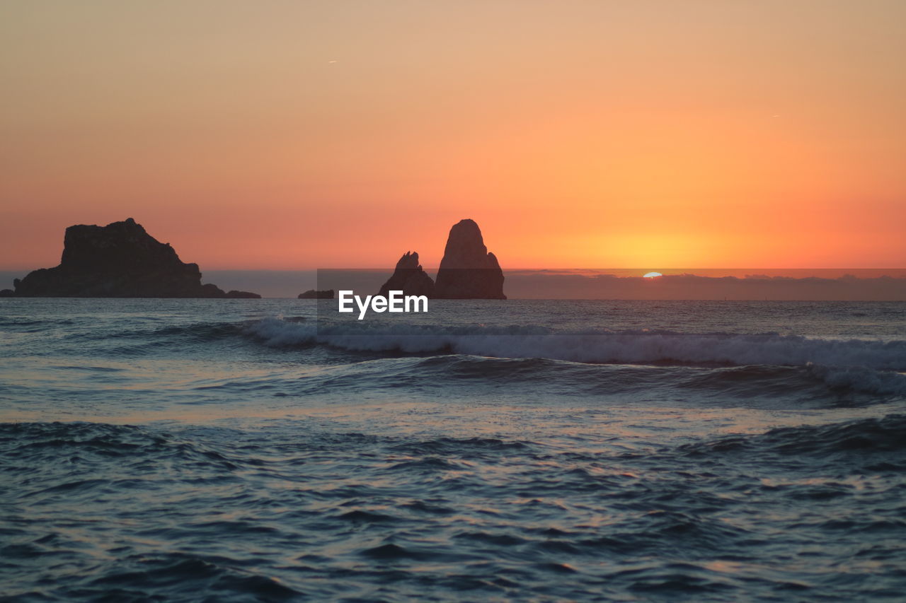 SCENIC VIEW OF ROCKS IN SEA AGAINST ORANGE SKY