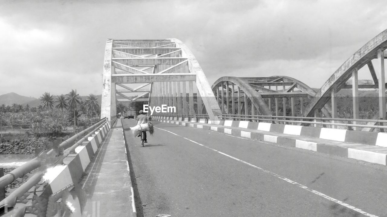 Rear view of man riding bicycle on bridge against cloudy sky