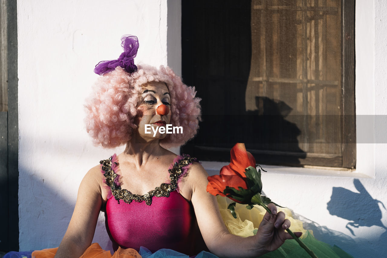 Serious mature woman with bright makeup in clown costume and wig looking at artificial red flower and looking away during performance on street