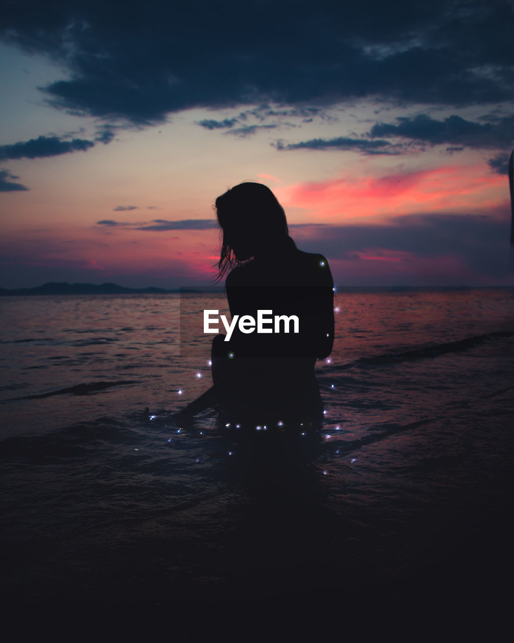 Silhouette woman with illuminated string lights in sea against sky during sunset