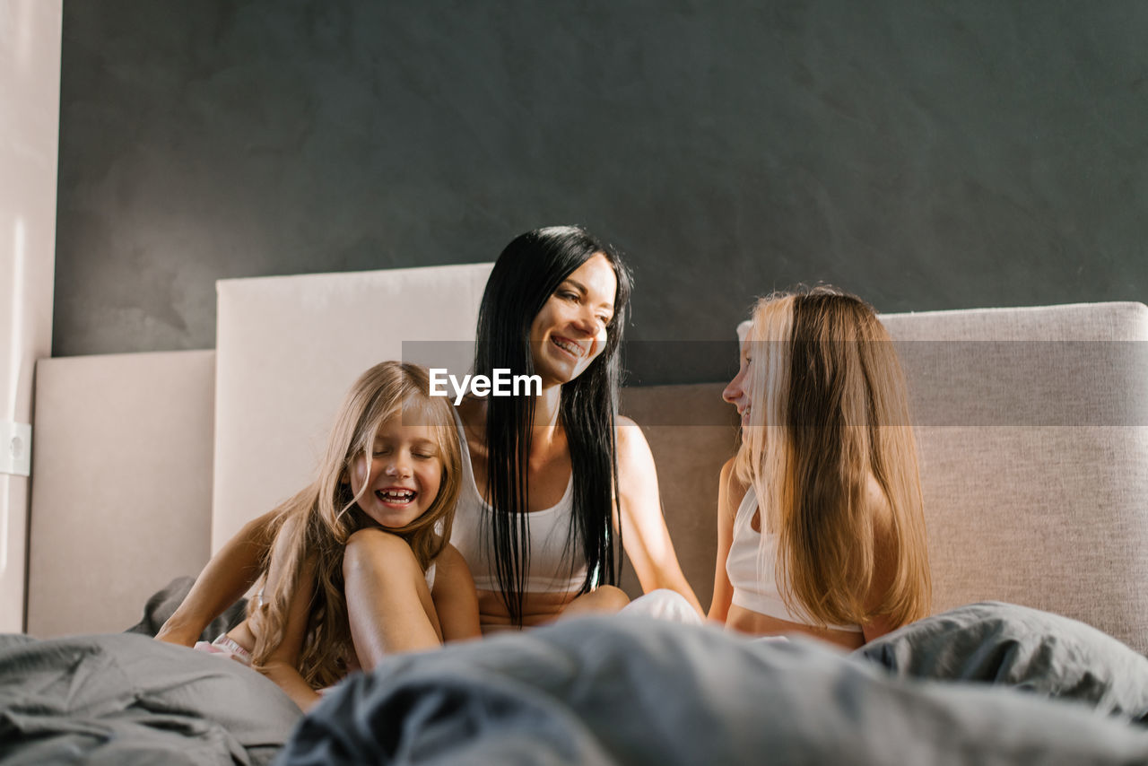 Young caucasian mother communicates with her daughters, sitting on the bed in the morning 