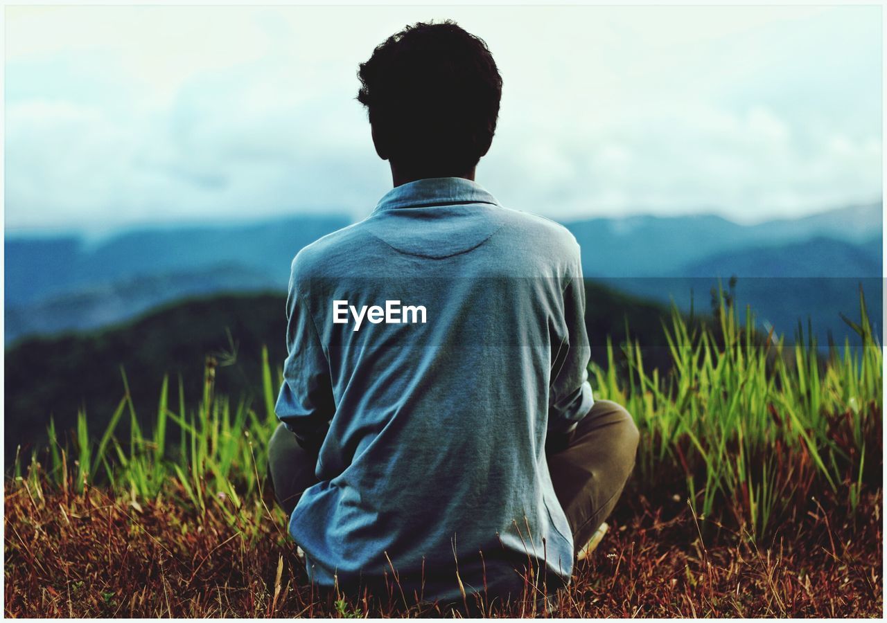 Rear view of young man sitting on field against sky