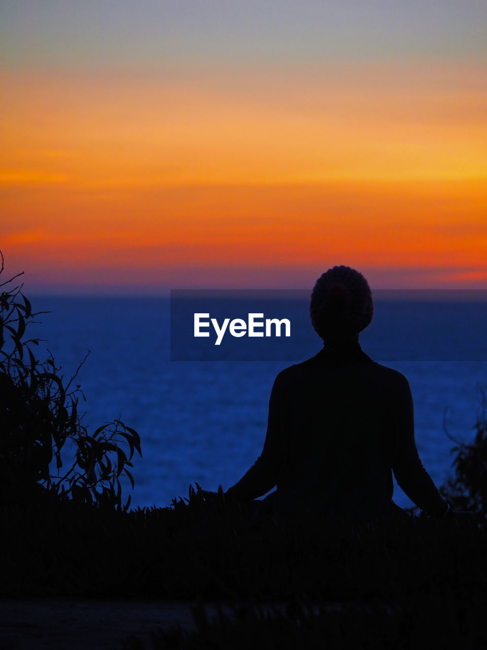 Silhouette man meditating in lotus position on cliff against sea during sunset