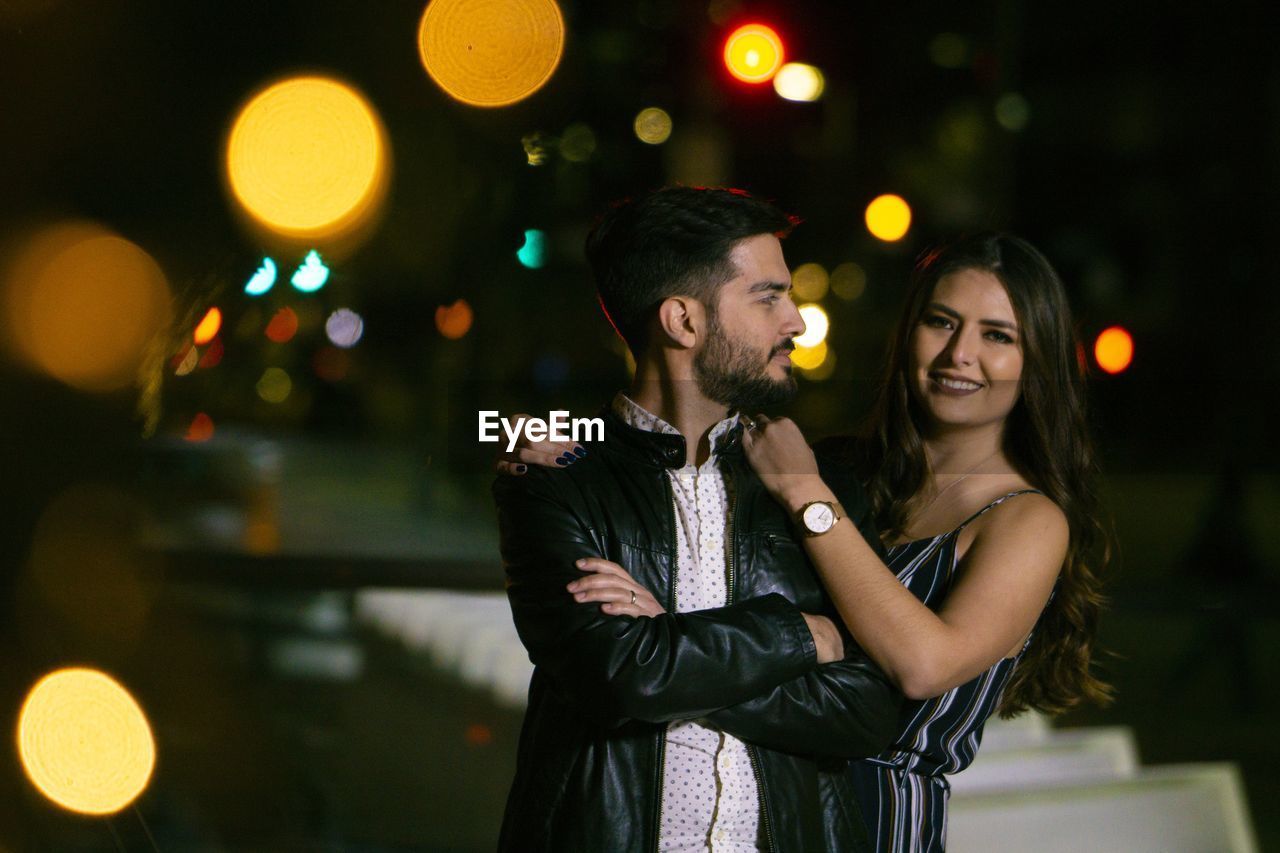 Portrait of smiling girlfriend holding boyfriend while standing in illuminated city at night