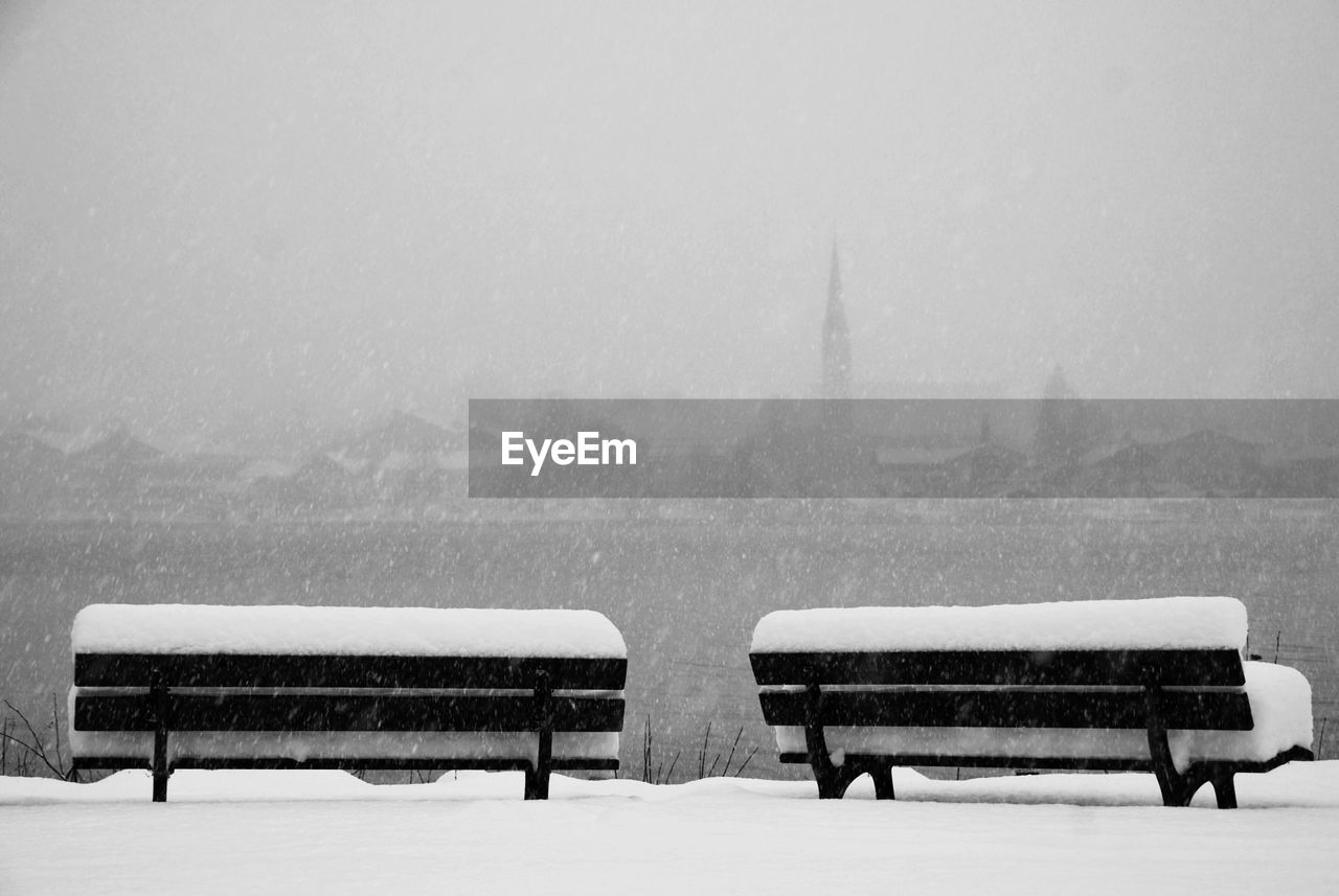 Snow covered benches against sky during snowfall