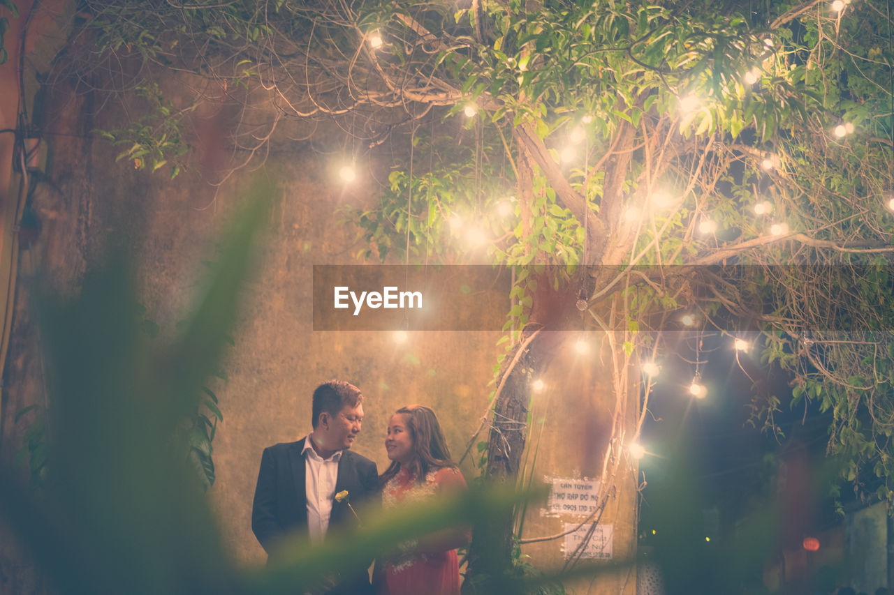 Couple standing below illuminated lighting decoration on tree at night