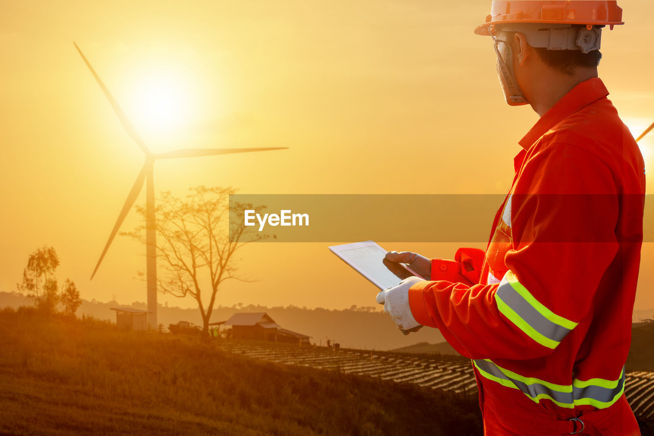 Civil engineer with digital tablet looking at windmill while standing against sky during sunset