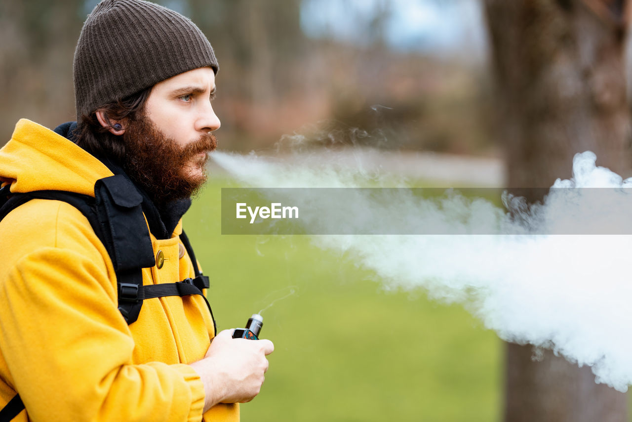 Side view of unshaven male traveler in outerwear with e liquid bottle vaping and looking forward on meadow