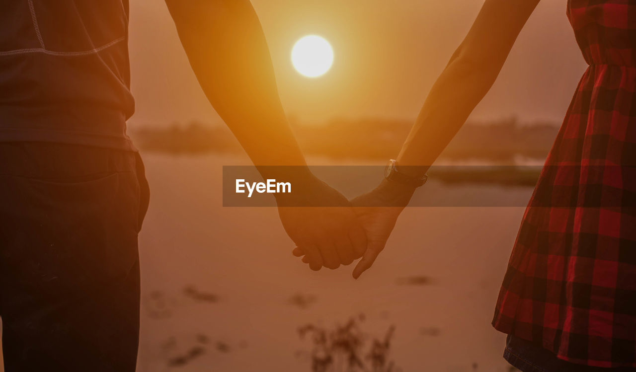 Midsection of couple holding hands while standing against sky during sunset