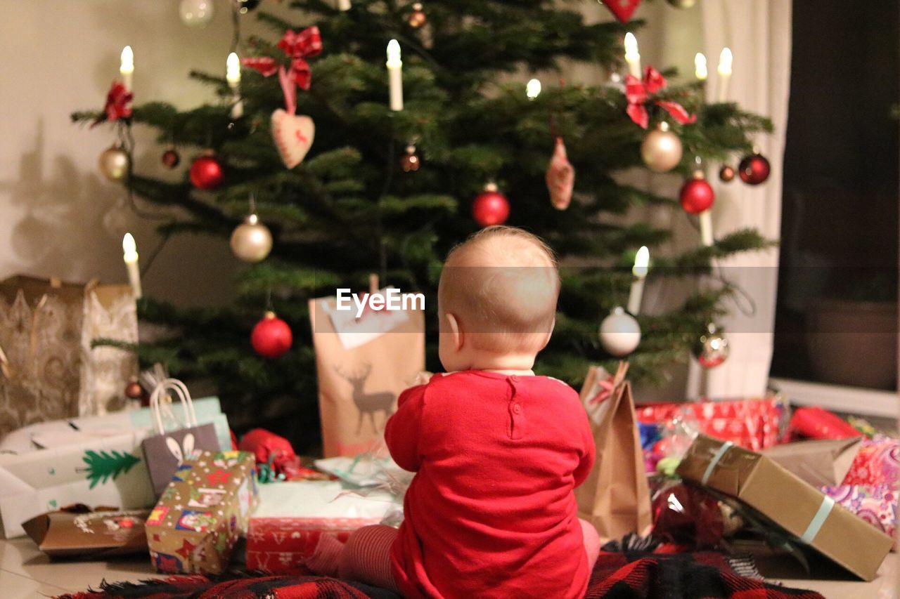 Rear view of baby sitting by gifts against christmas tree at home