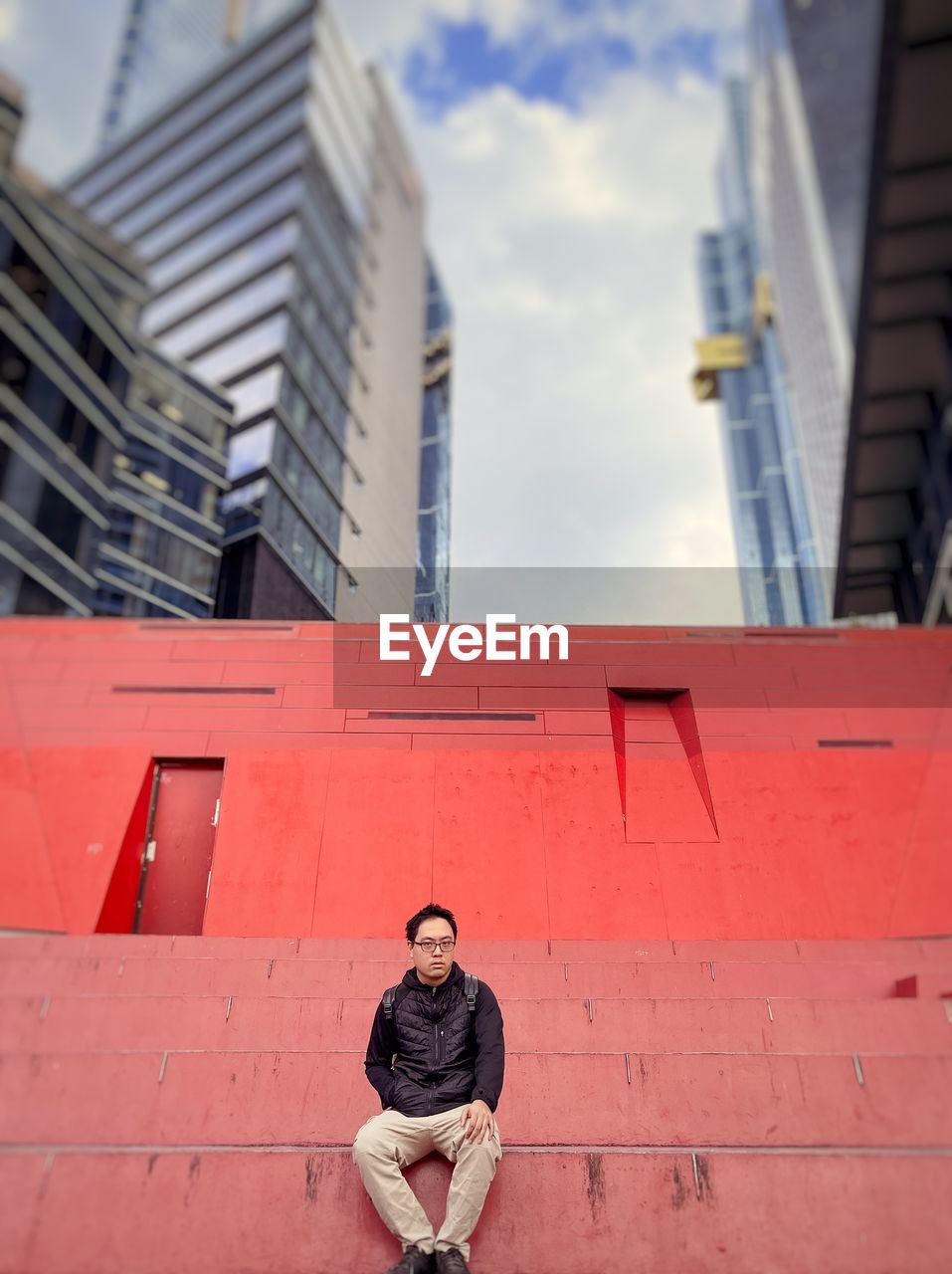 Low angle view of asian man sitting on steps against orange wall and skyscrapers in the city.