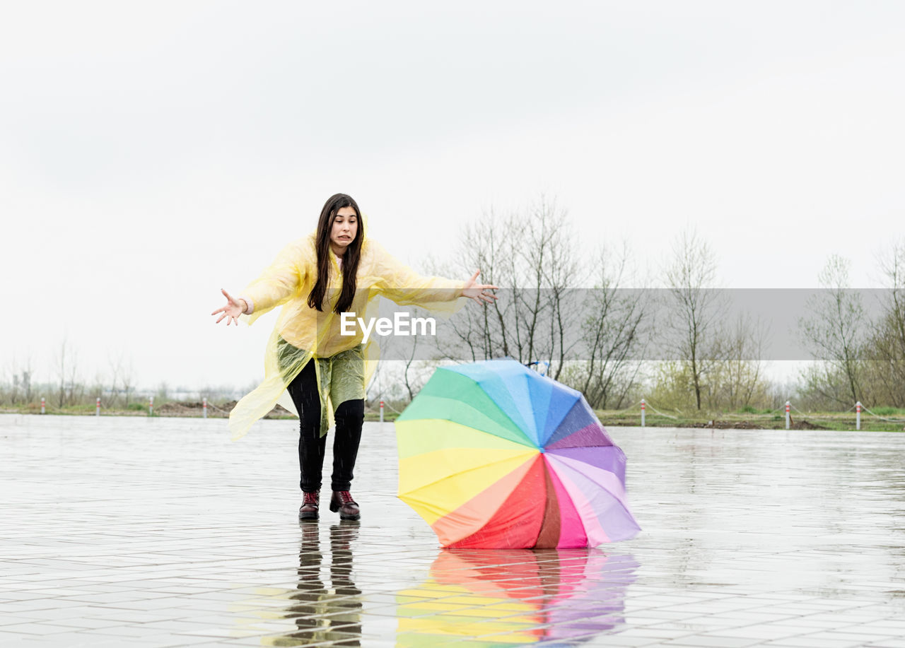 Beautiful brunette woman with funny face in yellow raincoat catching colorful umbrella