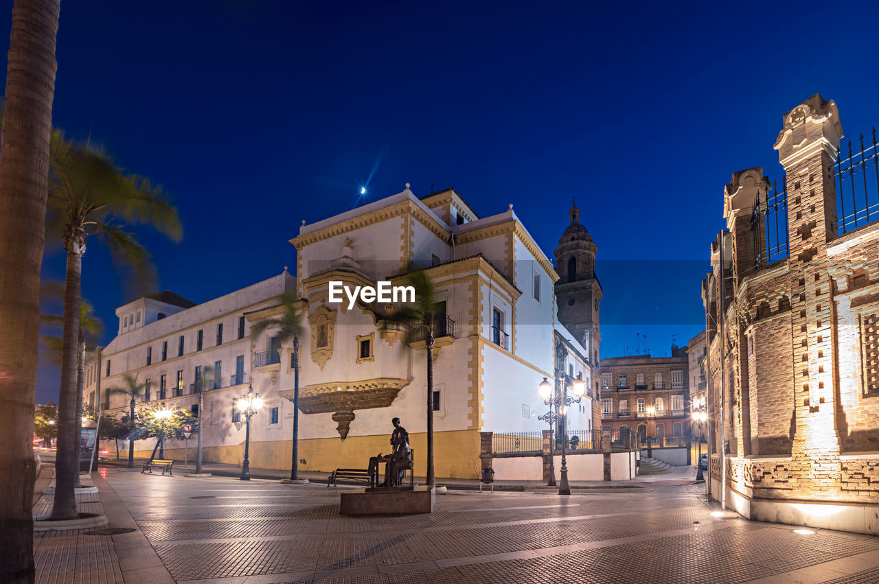 ILLUMINATED STREET AMIDST BUILDINGS AGAINST BLUE SKY