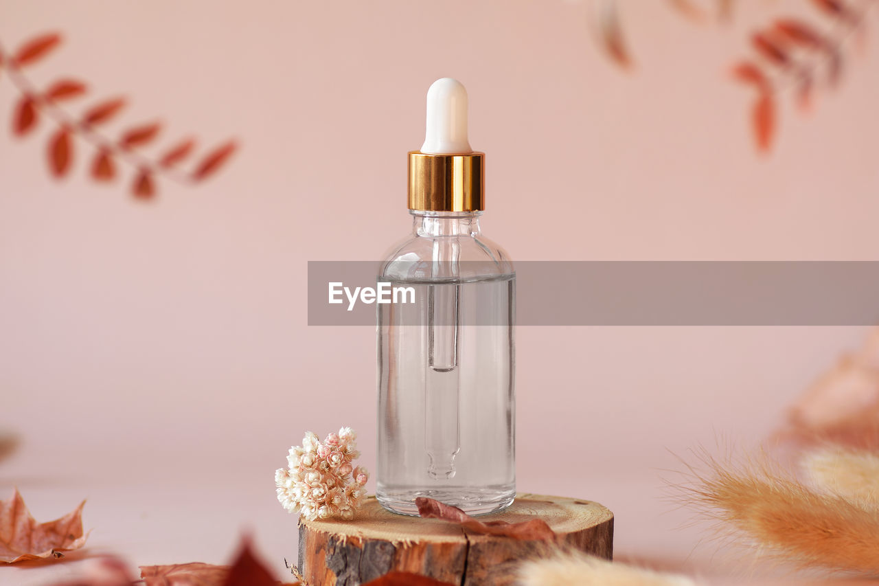 Transparent glass cosmetic dropper bottle standing on wooden disk with dry flowers and leaves