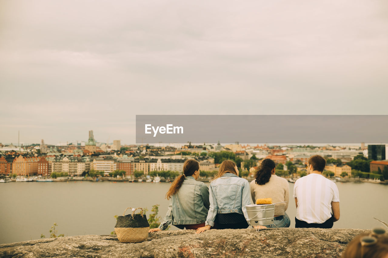 Rear view of friends sitting on rock formation at lakeshore during picnic against sky