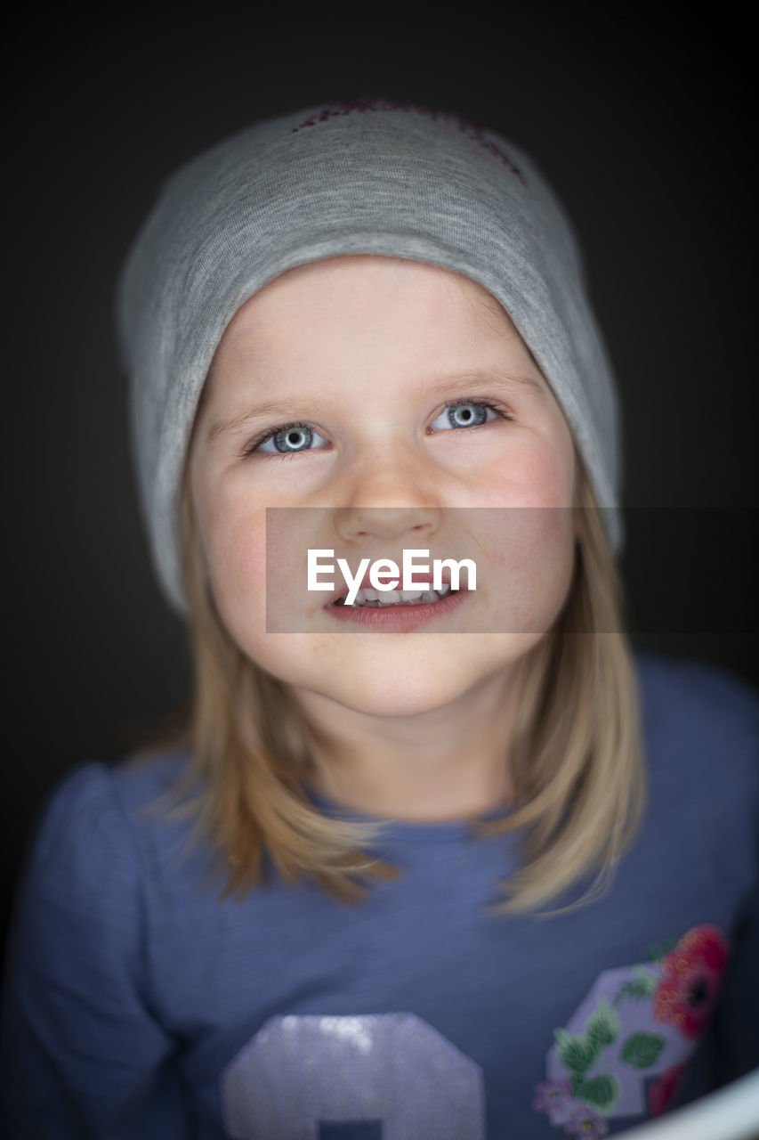 portrait, looking at camera, child, childhood, one person, front view, baby, skin, headshot, indoors, toddler, smiling, portrait photography, emotion, human face, black background, studio shot, happiness, women, close-up, clothing, innocence, blue, person, cap, female, human head, blond hair, cute, hat, casual clothing, white